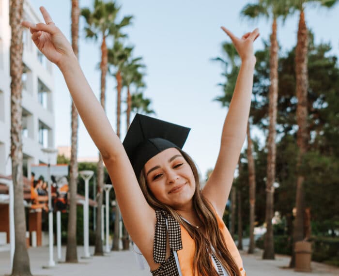 Outside a young graduate with mortar board is joyous with her arms in the air.